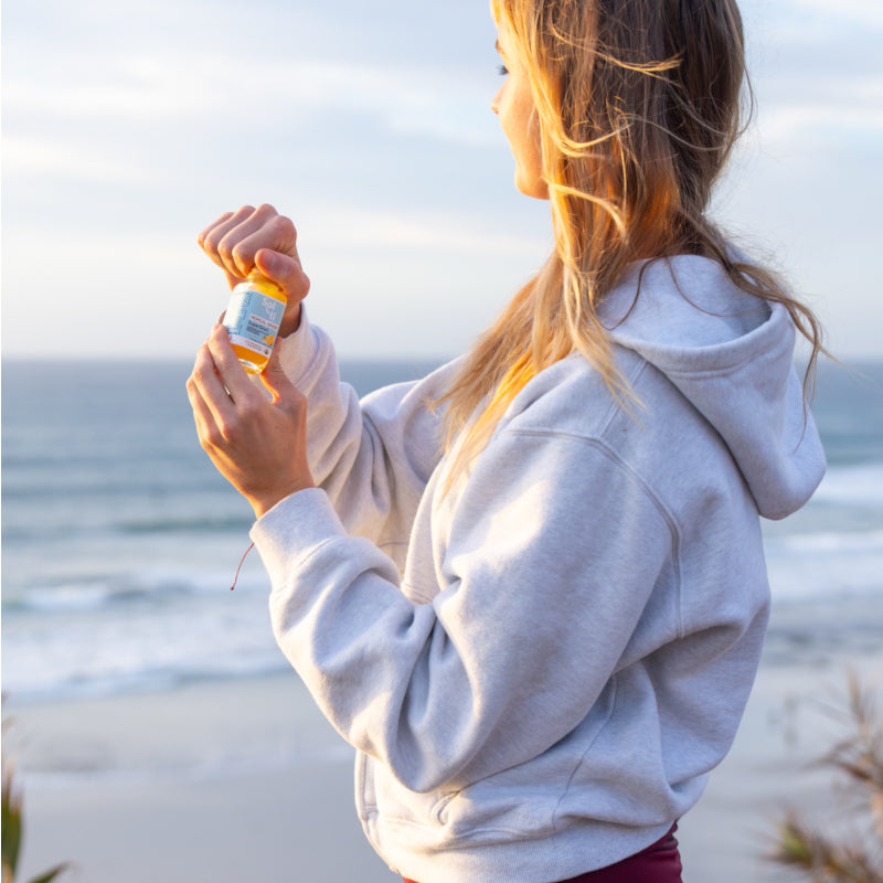 A young, woman with blonde hair in a light gray hoodie, holding a Sol-ti TROPICAL GINGER SuperShot, standing on a beach with waves and cloudy sky in the background.