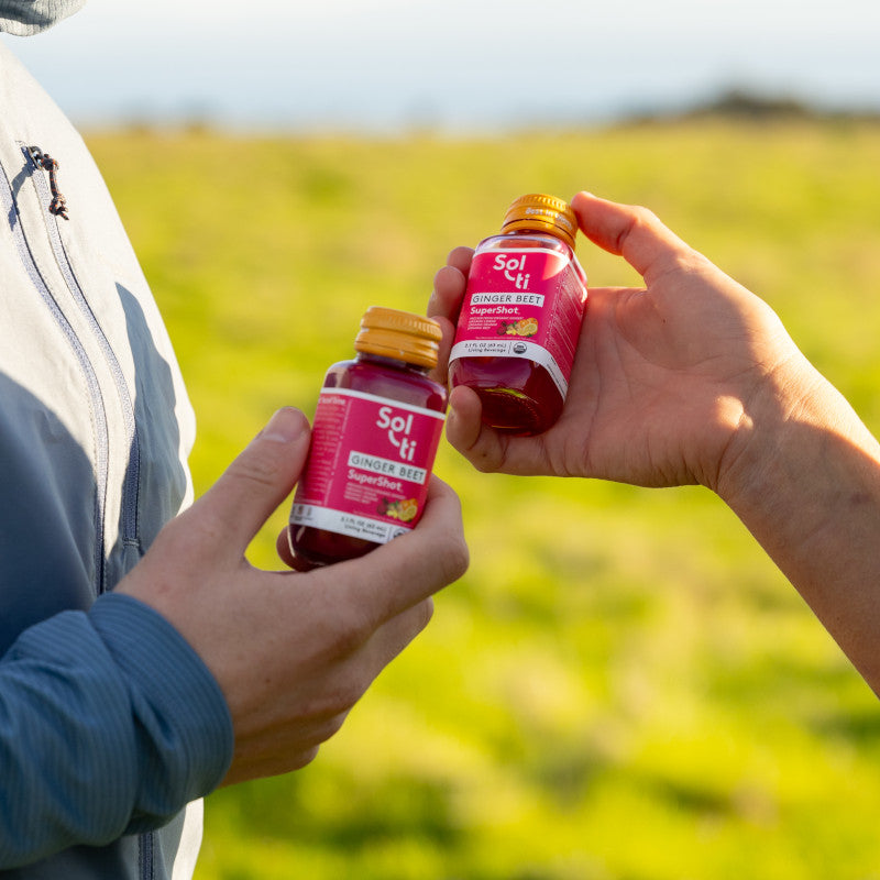 Two hands holding two bottles of Sol-ti GINGER BEET SuperShot, against a blurred green grassy background under bright sunlight.