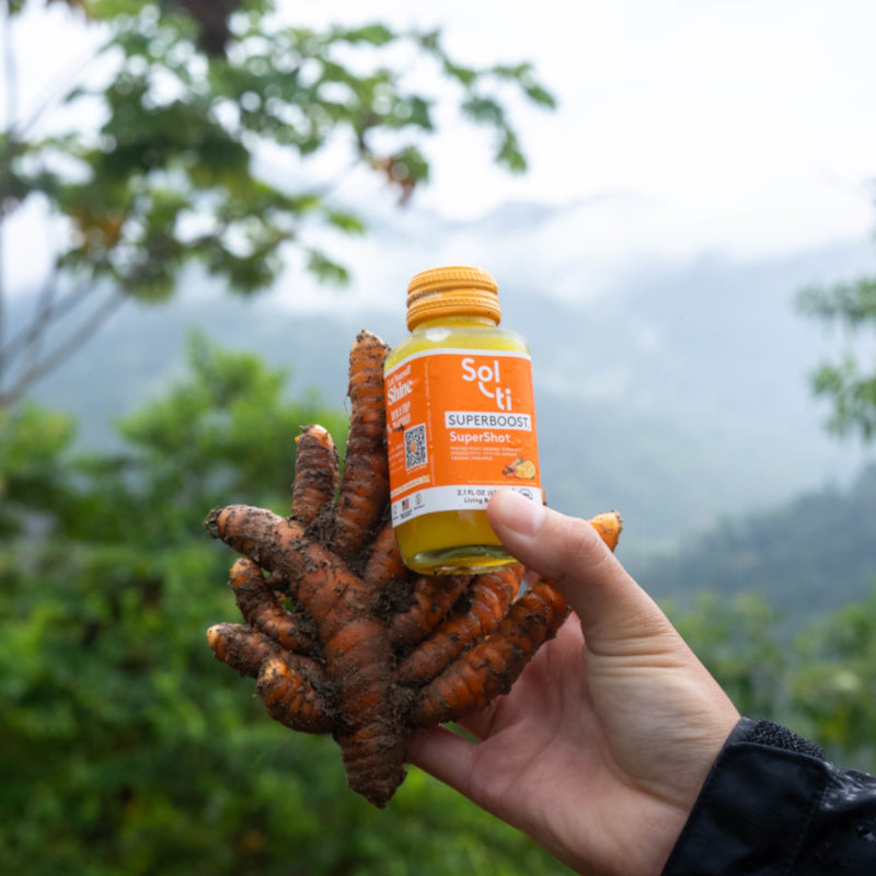 a hand holding a jar of SUPERBOOST SuperShot, with a fresh turmeric root in the background.