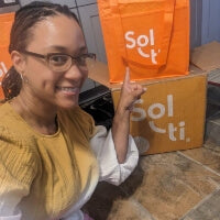 A woman smiling and sitting on the floor pointing up a Sol-ti bag with products