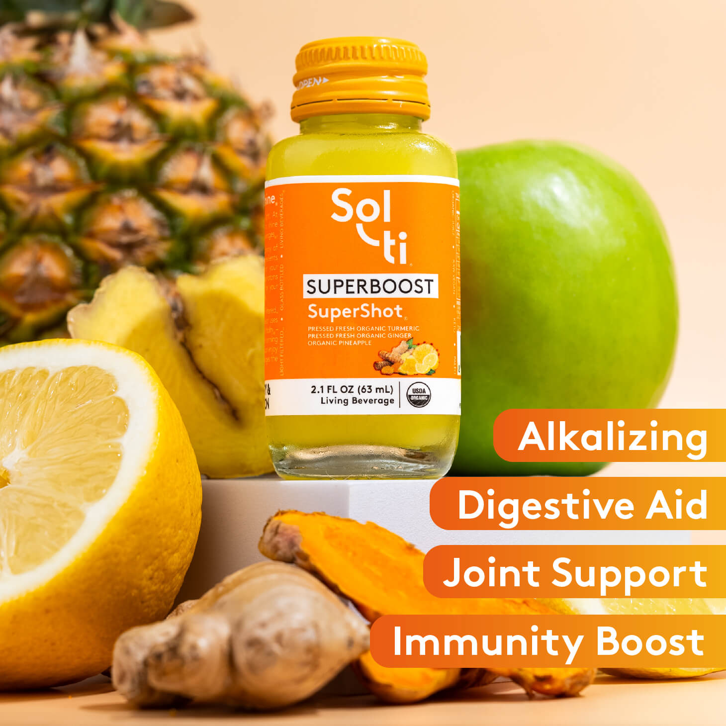 Sol-ti SUPERBOOST SuperShot glass bottle with front label on light orange background, with turmeric root, ginger root, pineapple, lemon slice and green apple, benefits text including Alkalizing, Digestive aid, Joint support and immunity boost