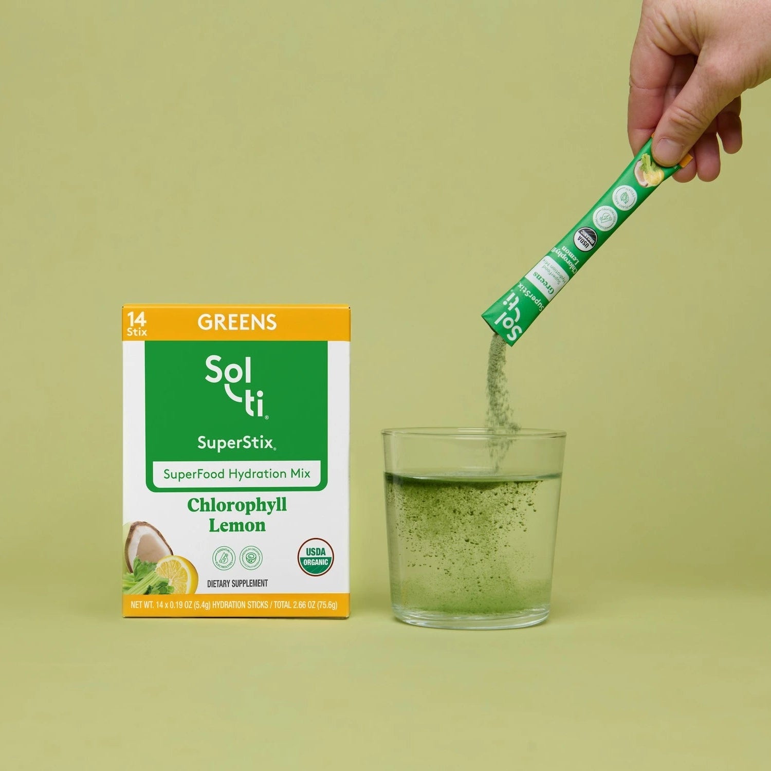 A hand pours green powder from a Sol-ti SuperStix Chlorophyll Lemon packet into a clear glass on a light olive-green background, with the Sol-ti greens box standing beside it.