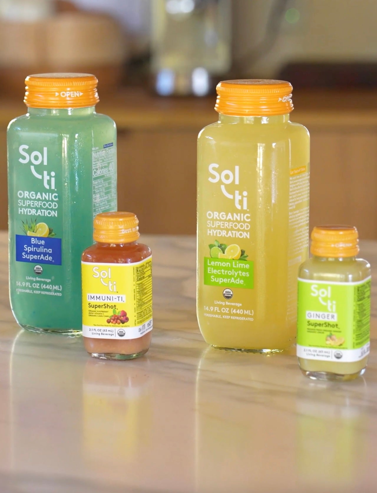 Assortment of Sol-ti organic bottled drinks on a kitchen counter, featuring two large SuperAde bottles (blue spirulina and lemon lime) and three smaller wellness shots (turmeric, IM-MUNI-TI, and ginger) with orange caps.