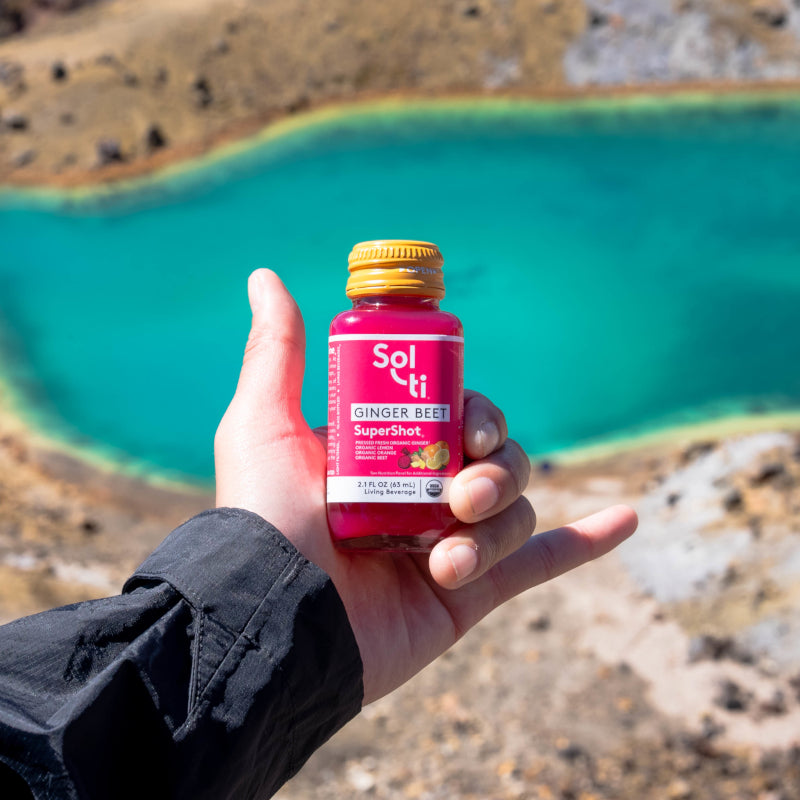 A hand holding a GINGER BEET SuperShot bottle with a yellow cap, against a backdrop of a turquoise hot spring surrounded by rocky, brown terrain.