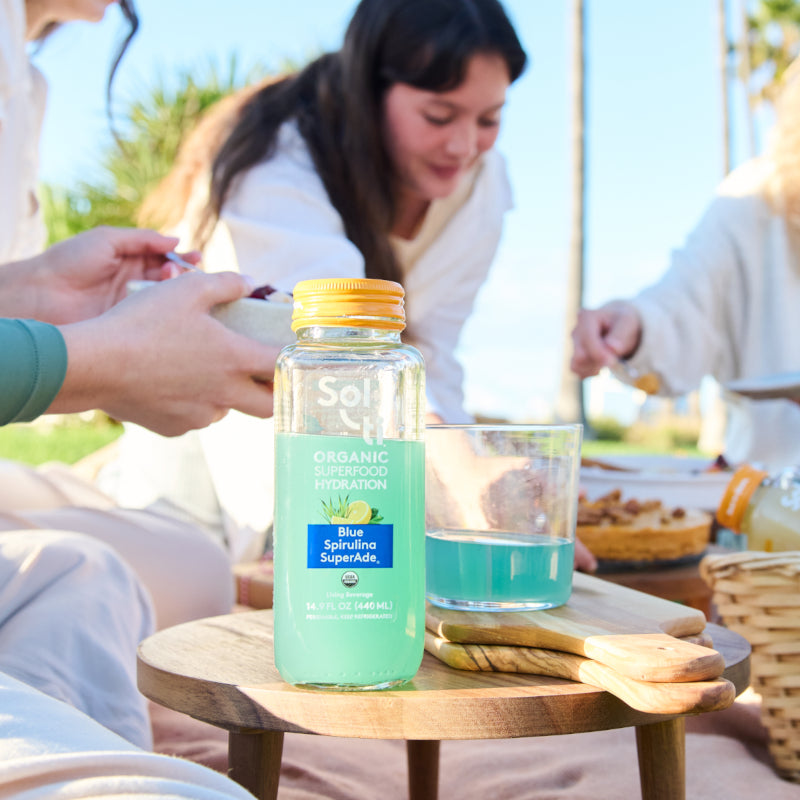 Sol-ti Blue Spirulina SuperAde bottle on table at outdoor gathering, front label, light background