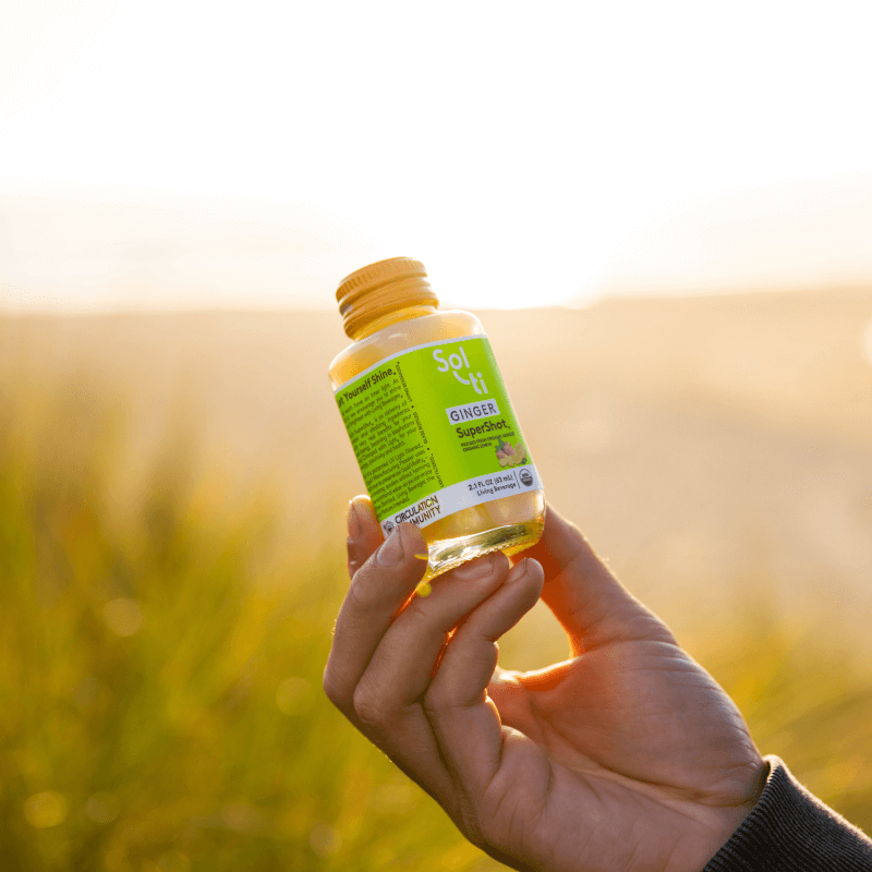 A hand holding a GINGER SuperShot, set against a blurred, sunlit grassy background, suggesting an outdoor, natural setting.