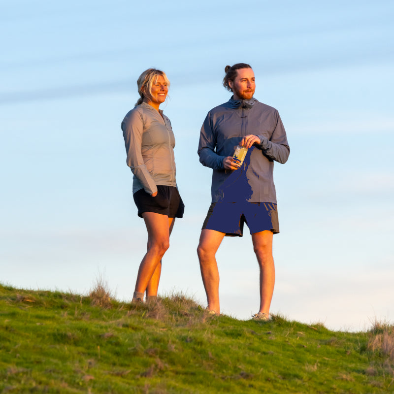 Two individuals stand on a grassy hilltop, after a run, pausing to take a break and enjoying the view with Lemon Lime SuperAde.