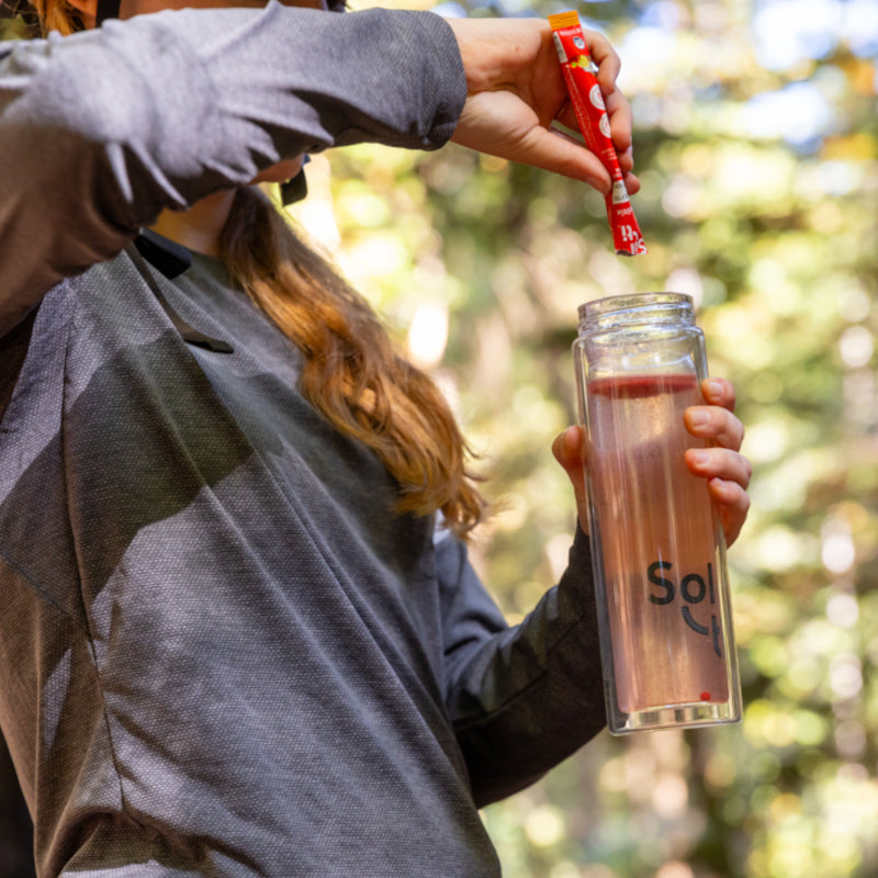 A woman wearing a gray long-sleeve shirt is pouring a red SuperStix mix from a small packet into a clear water bottle outdoors. 