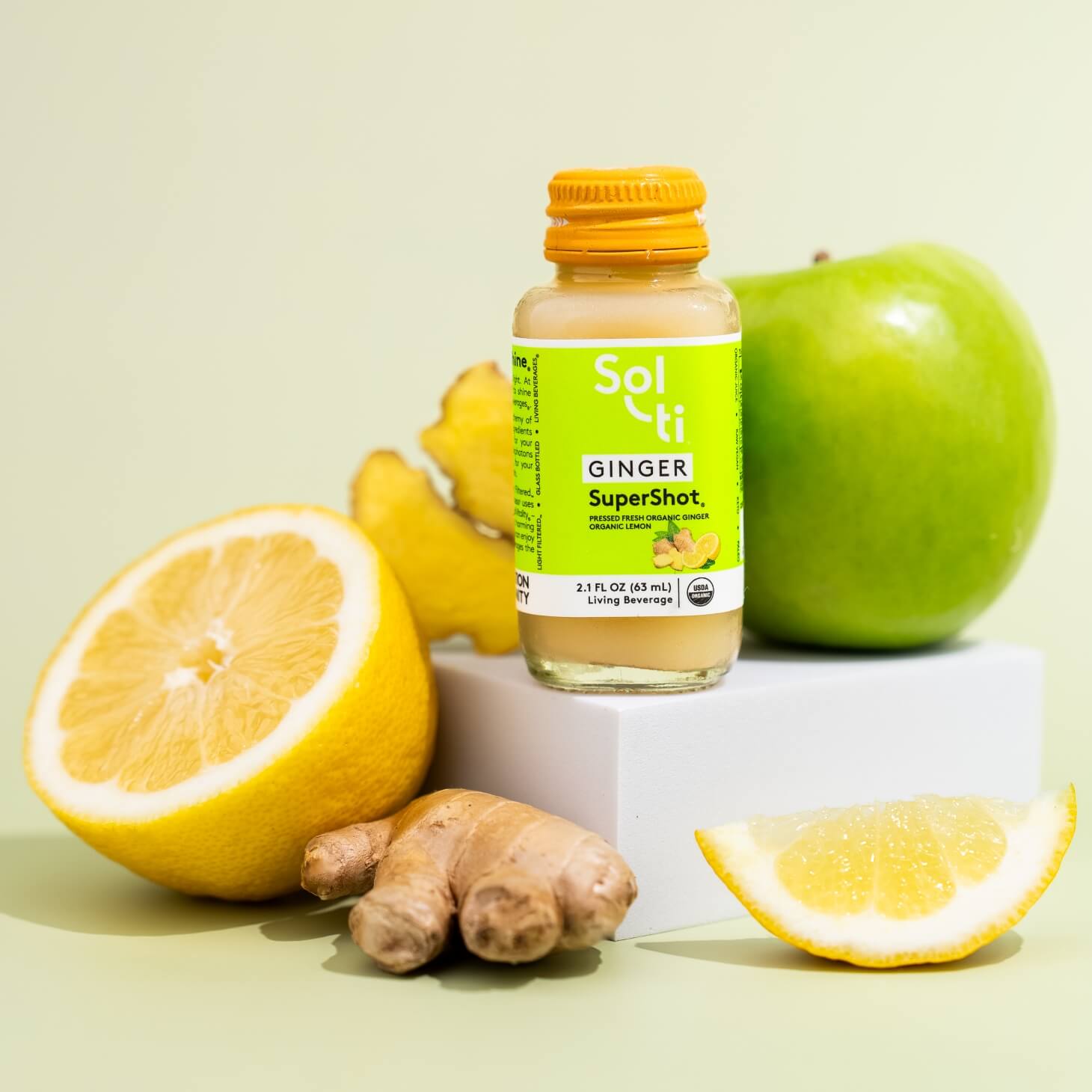 Sol-ti GINGER SuperShot bottle, green apple, lemon, ginger root, and lime, all on a white block against a light green background.