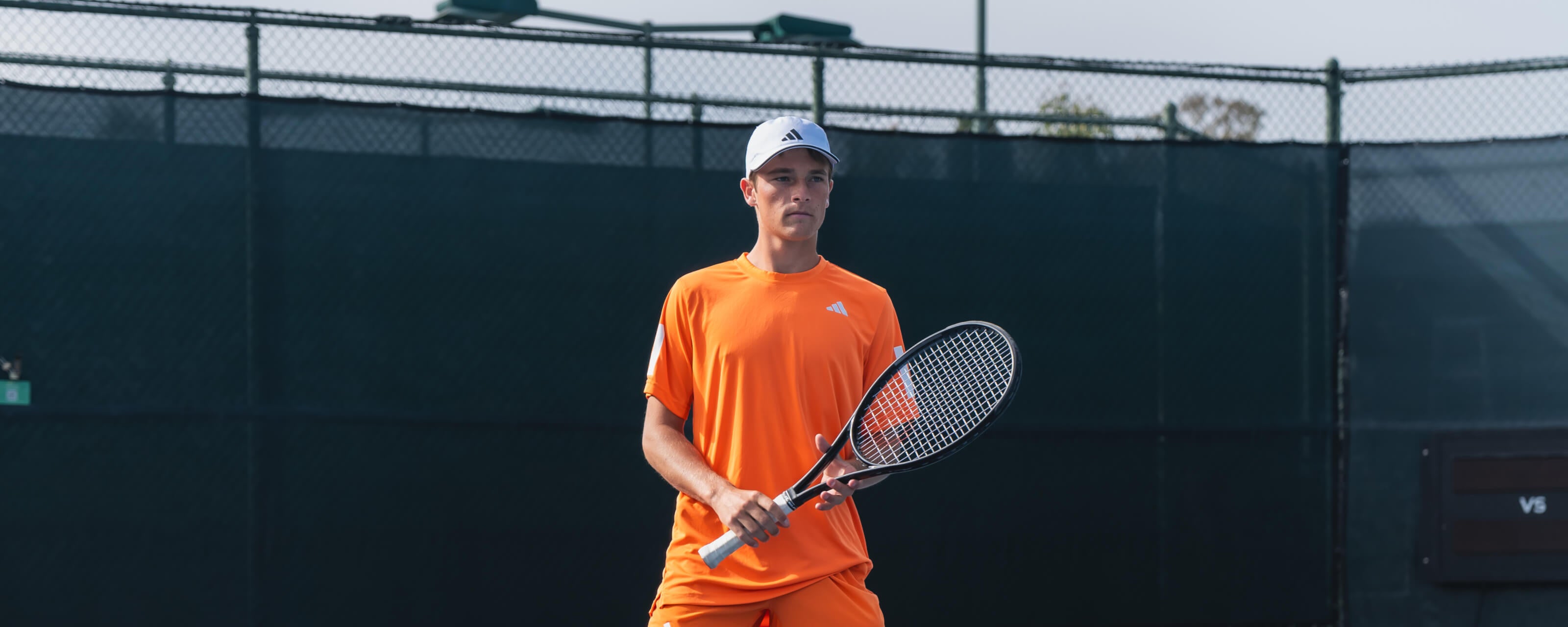 Andy Johnson holding a tennis racket stands on a blue hard court in front of a dark fence