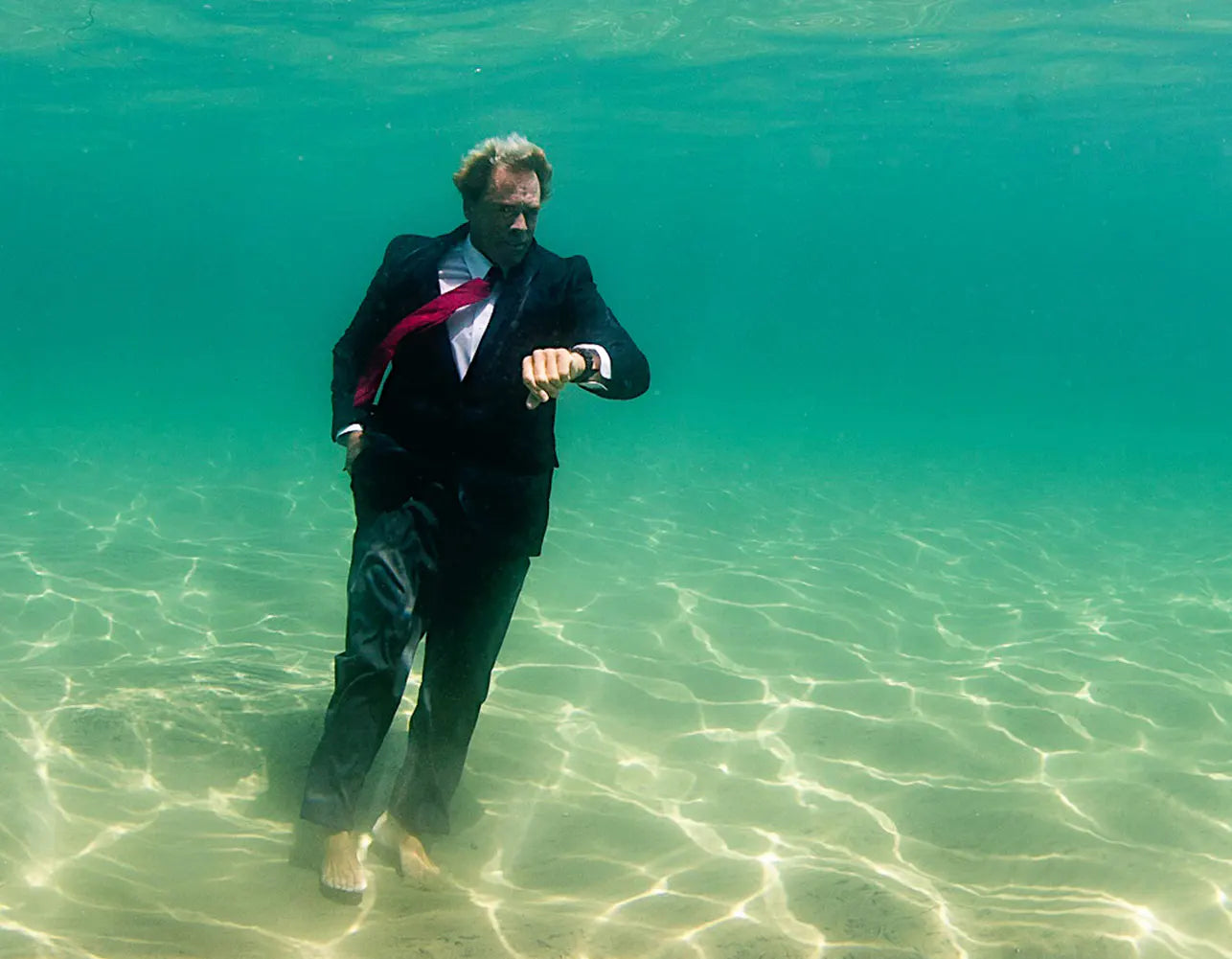 Chuck Patterson in a black suit stands barefoot underwater in clear turquoise water
