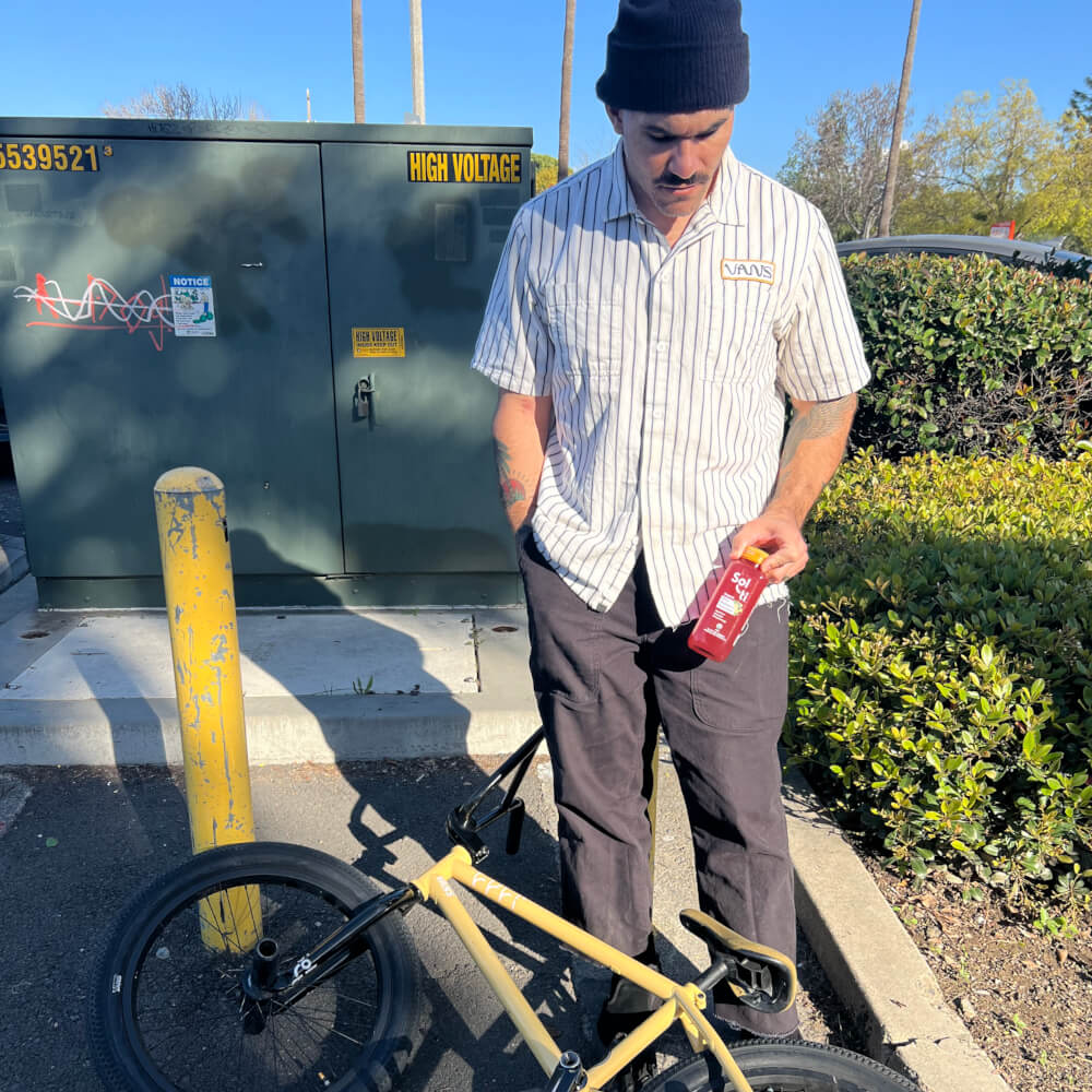 Dakota Roche stands next to a yellow BMX bike on an outdoor area, holding a Wild Blueberry SuperAde