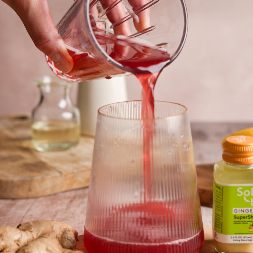 Kombucha being poured from a glass container into another glass on a wooden surface with a bottle of Sol-ti GINGER SuperShot in the background.