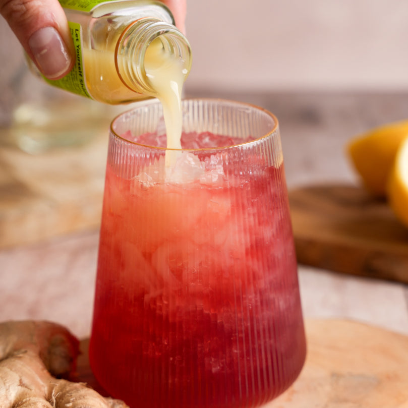Person pouring GINGER SuperShot into a glass of Kombucha with ice, surrounded by ginger and lemons.