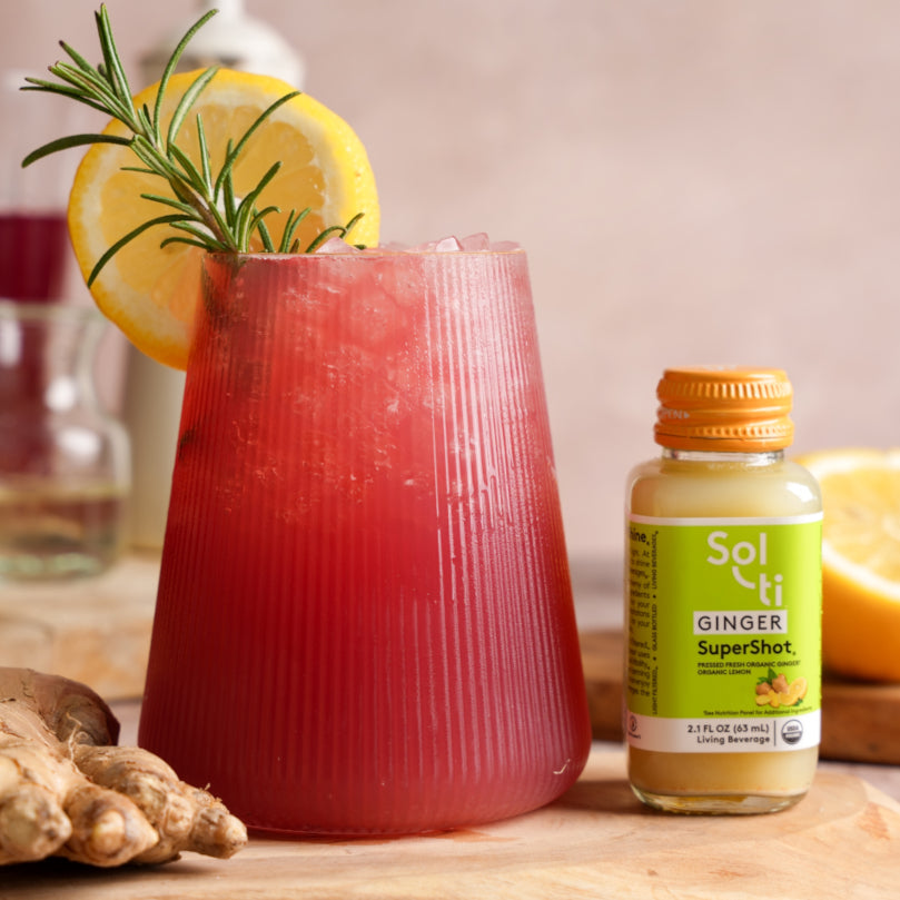 Digestive Aid Mocktail in a glass with lemon and rosemary, next to a bottle of Sol-ti GINGER SuperShot on a wooden surface.