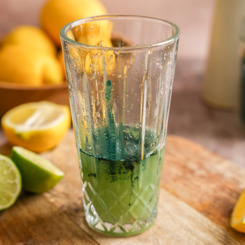 Clear glass shaker with green spirulina and lemin on a wooden surface with lemons and limes.