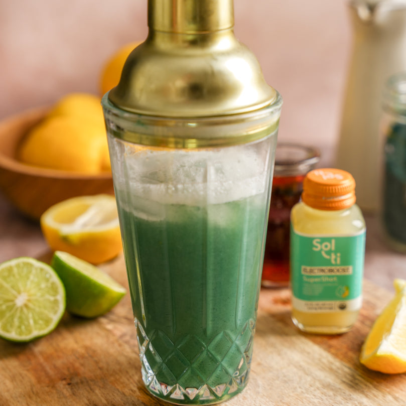 Green spirulina lemonada in a shaker with a gold lid, surrounded by lemons, limes, and a bottle of ELECTROBOOST SuperShot on a wooden surface.