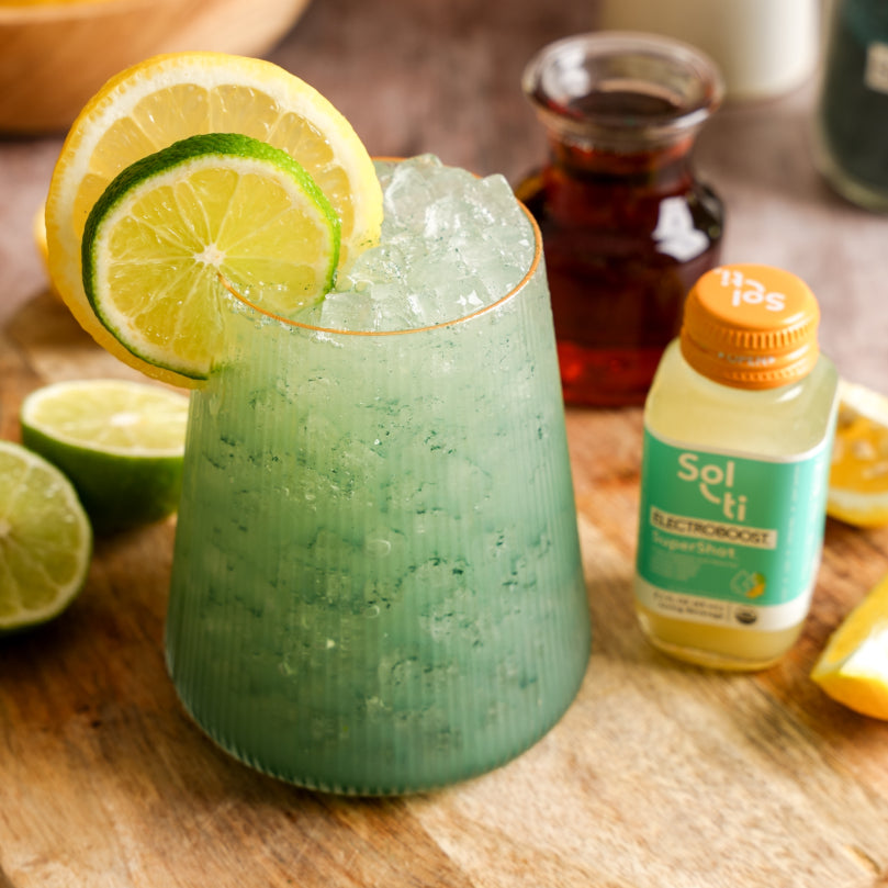 Electroboost Green Spirulina Lemonade drink with lemon and lime slices on a wooden surface, with Sol-ti ELECTROBOOST™ SuperShot glass bottle