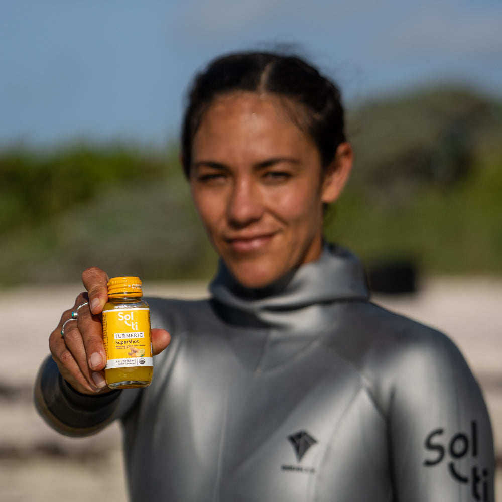 Enchante Gallardo in a gray wetsuit holds a TURMERIC SuperShoti on a beach