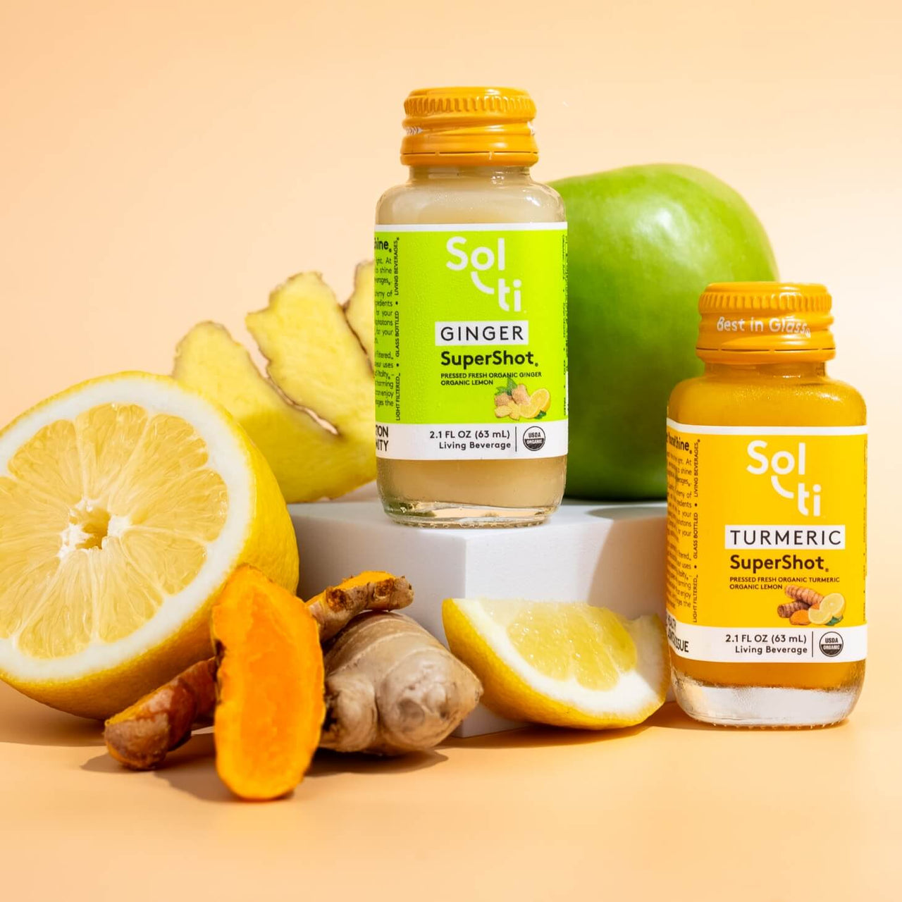 a bottle of GINGER SuperShot next to a lemon, ginger root and a apple, a TURMERIC SuperShot bottle with an apple, turmeric root  and a lemon slice 