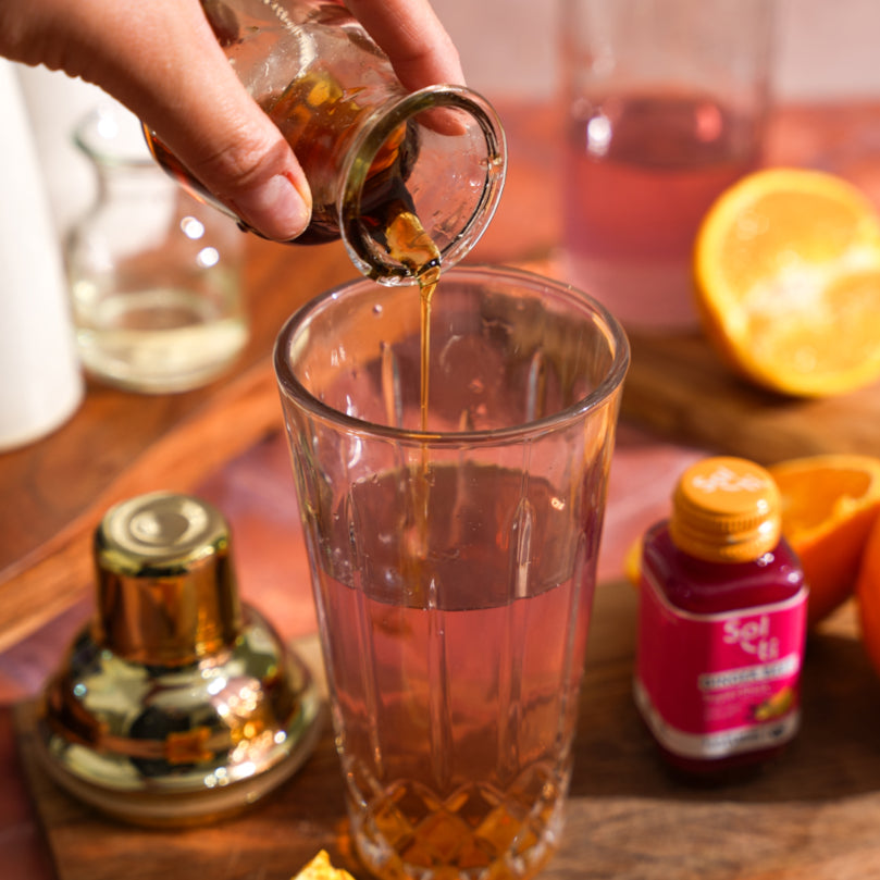 Hand pouring Maple Syrup into a glass on a wooden surface with oranges and other items.