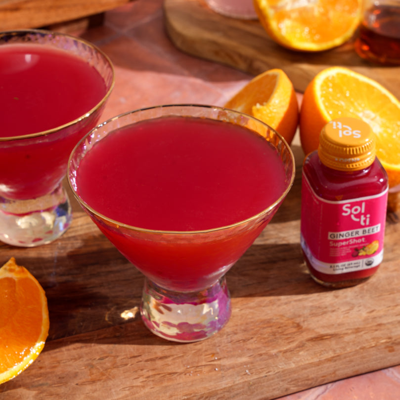 Two glasses of Ginger Beet Recovery Drink with a bottle of Sol-ti GINGER BEET SuperShot® and orange slices on a wooden surface.