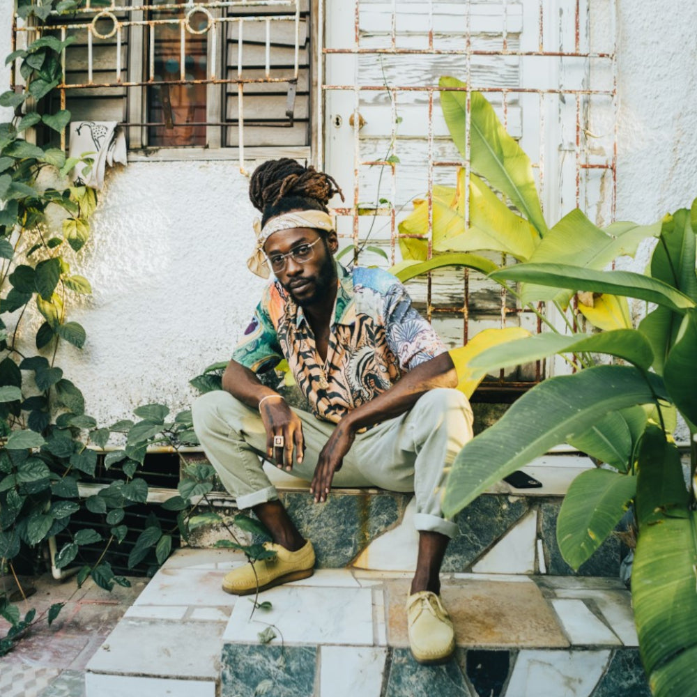 Jesse Royal sits on tiled steps in front of a white wall with plants and a metal barred window