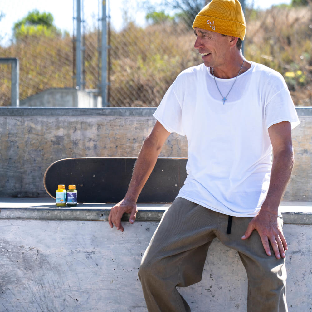 Joey Brenzinski sits on a concrete skatepark ledge with a skateboard and  two SuperShot bottles
