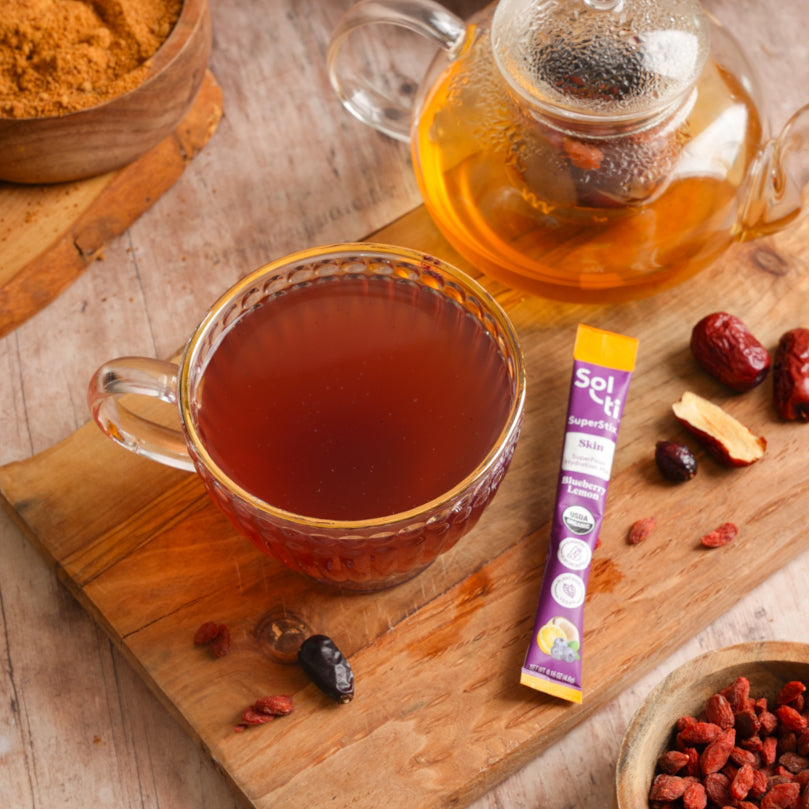 Clear mug of tea with a teapot, jujubes, and a Sol-ti Blueberry Lemon SuperStix on a wooden surface