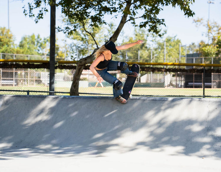 Katelyn West is performing a skateboard trick on a concrete ramp at an outdoor skatepark