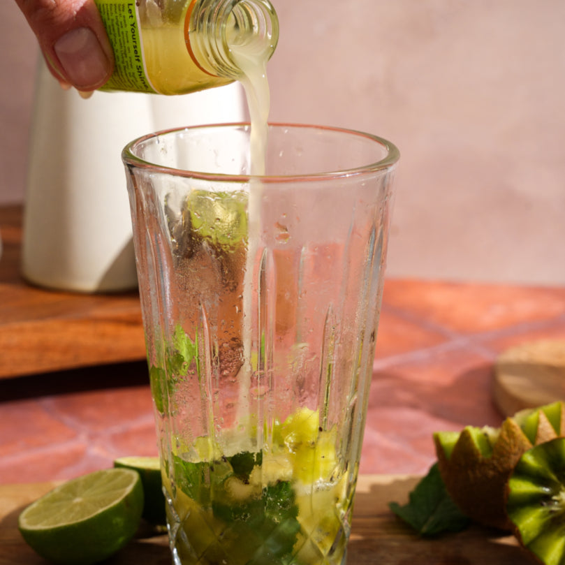 Hand pouring GINGER SuperShot® into a glass with kiwi, mint leaves, and lime juice on a wooden surface.