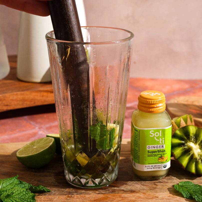 Muddling ingredients in a glass with a bottle of Sol-ti GINGER SuperShot® on a wooden surface.