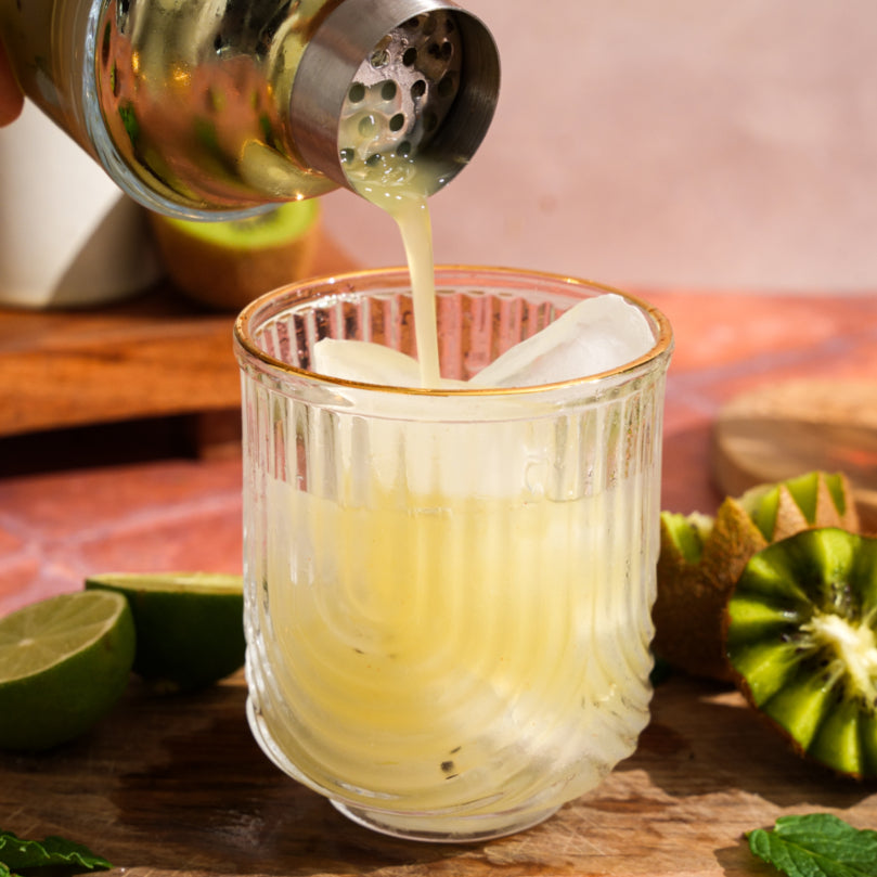 Kiwi Ginger Mojito Mocktail being poured into a glass with lime and kiwi on a wooden surface.