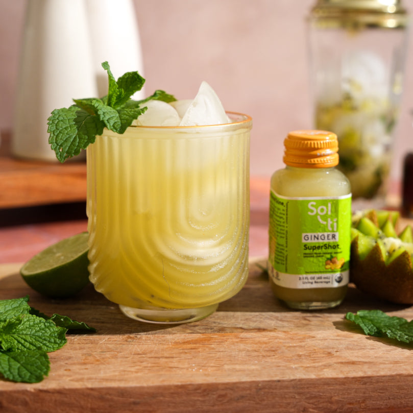 Glass of Kiwi Ginger Mojito Mocktail with ice and mint levaes, Sol-ti GINGER SuperShot® bottle, on a wooden surface.