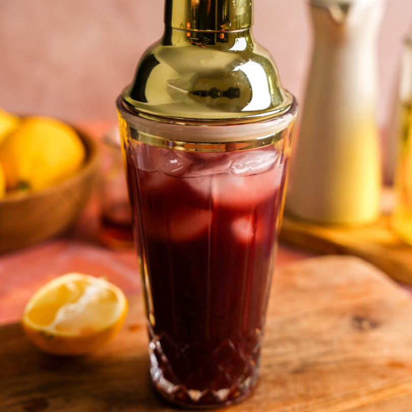 Shaker with red liquid and ice cubes on a wooden surface with lemons in the background