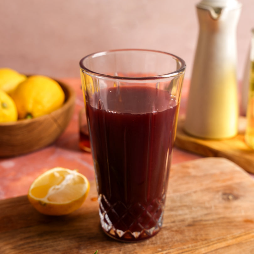 Glass shaker with dark red liquid on a wooden surface with lemons and a pitcher in the background.