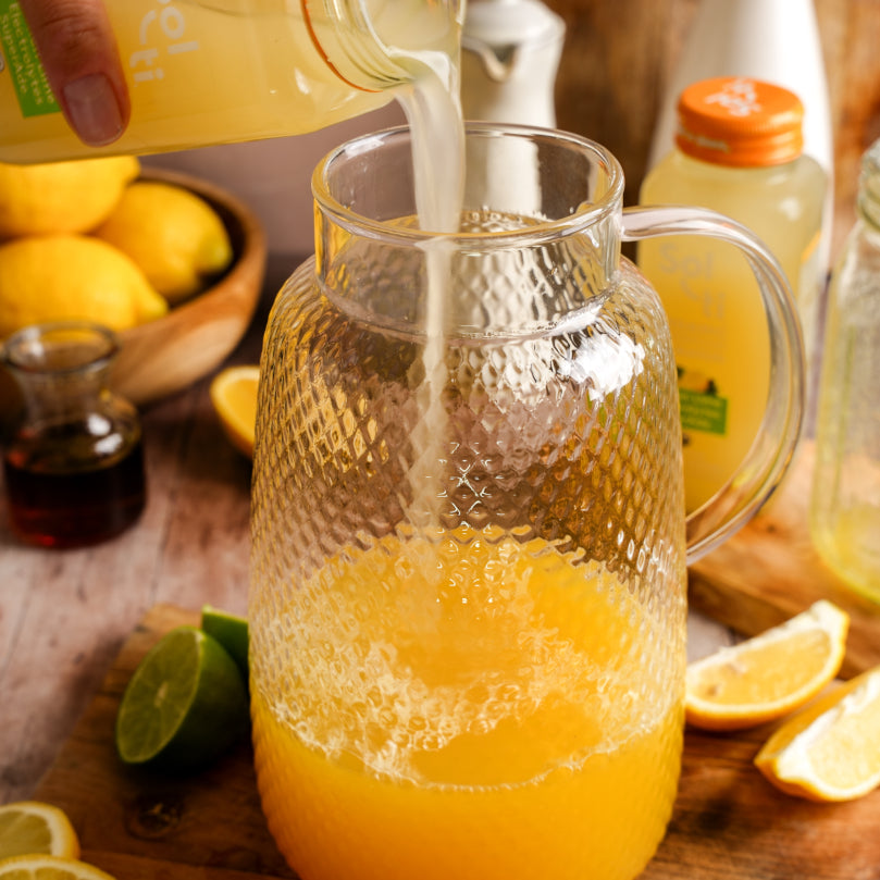 Pineapple juice and Lemon Lime SuperAde being poured into a glass pitcher with lemons and limes on a wooden surface.