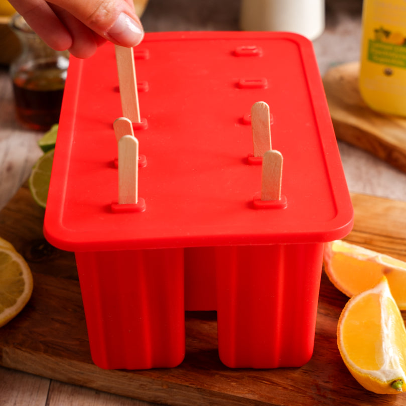 Red silicone popsicle mold with wooden sticks on a wooden surface with lemons and a Lemon Lime SuperAde glass bottle in the background.
