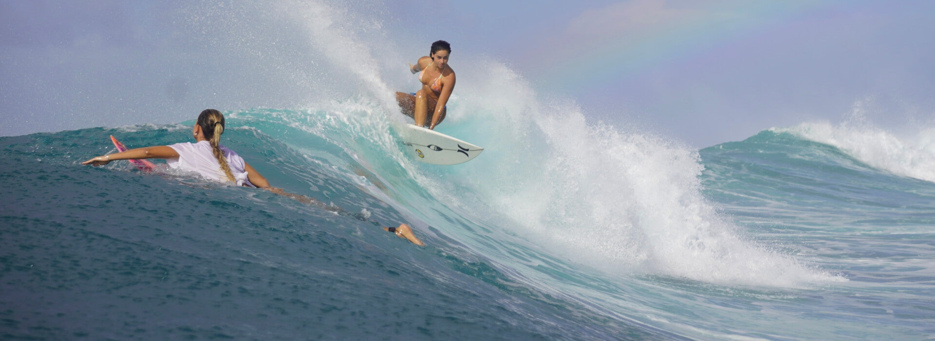 Molly Tuschen stands on a surfboard riding a turquoise ocean wave under a pale blue sky