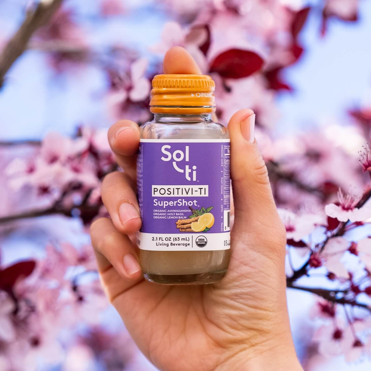 a hand holding a bottle of POSITIVI-TI SuperShot in front of cherry blossom flowers