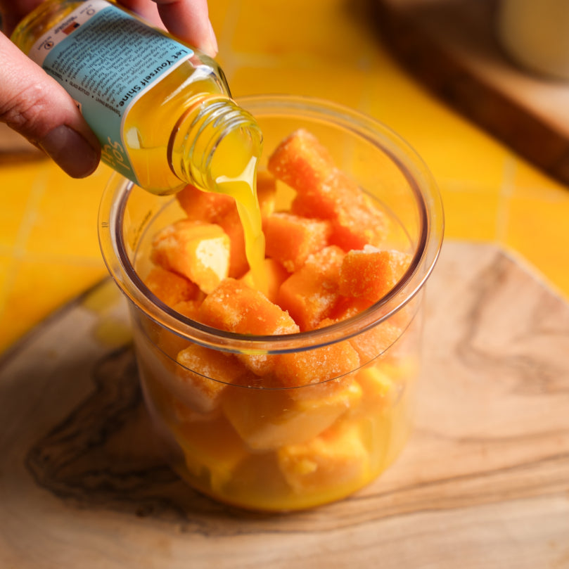 Hand pouring a TROPICAL GINGER SuperShot from a bottle into a container of chopped ffrozen mango and frozen passion fruit on a wooden surface.