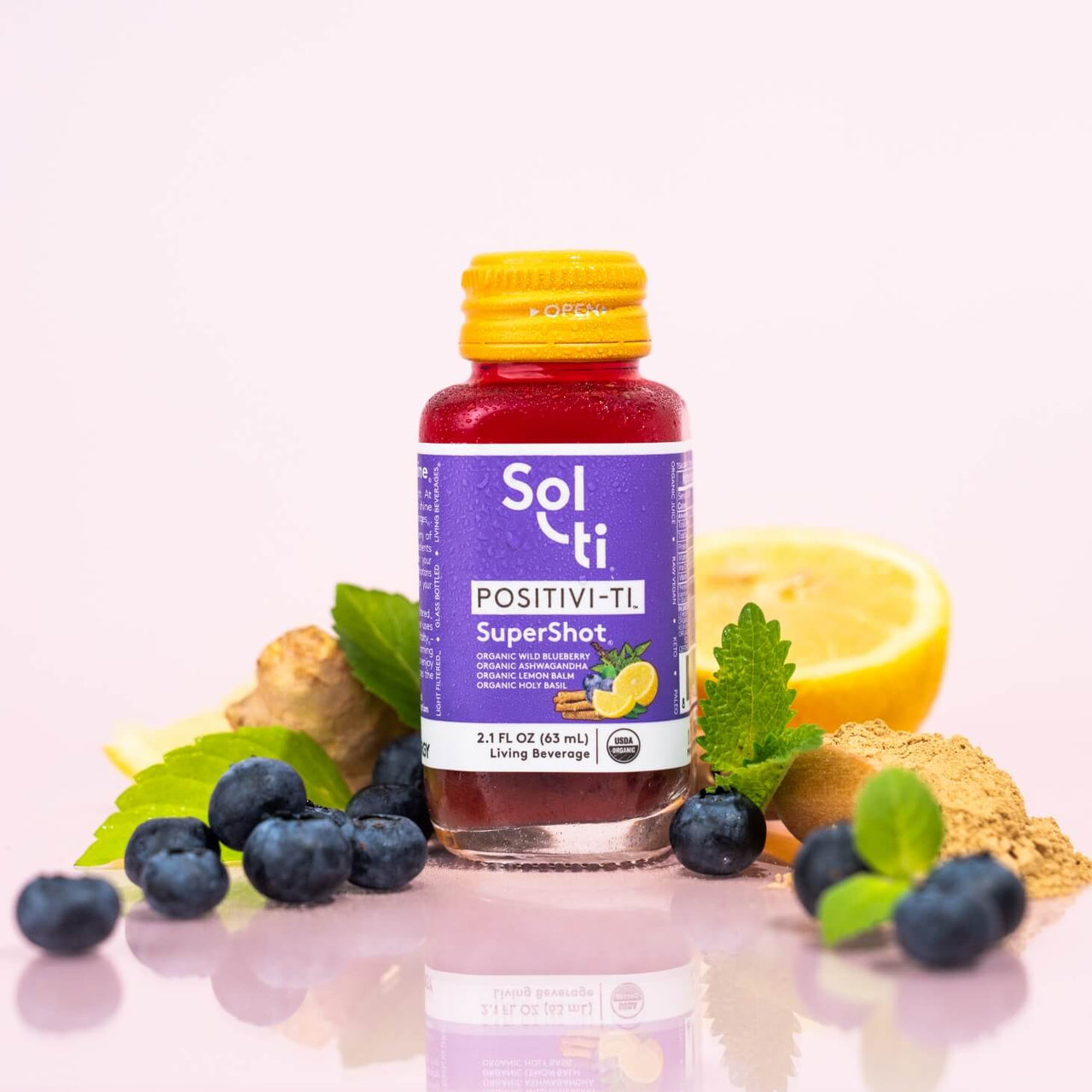 A bottle of POSITIVI-TI SuperShot next to Ashwagandha root, wild blueberries, lemon balm leaves, and lemon slices