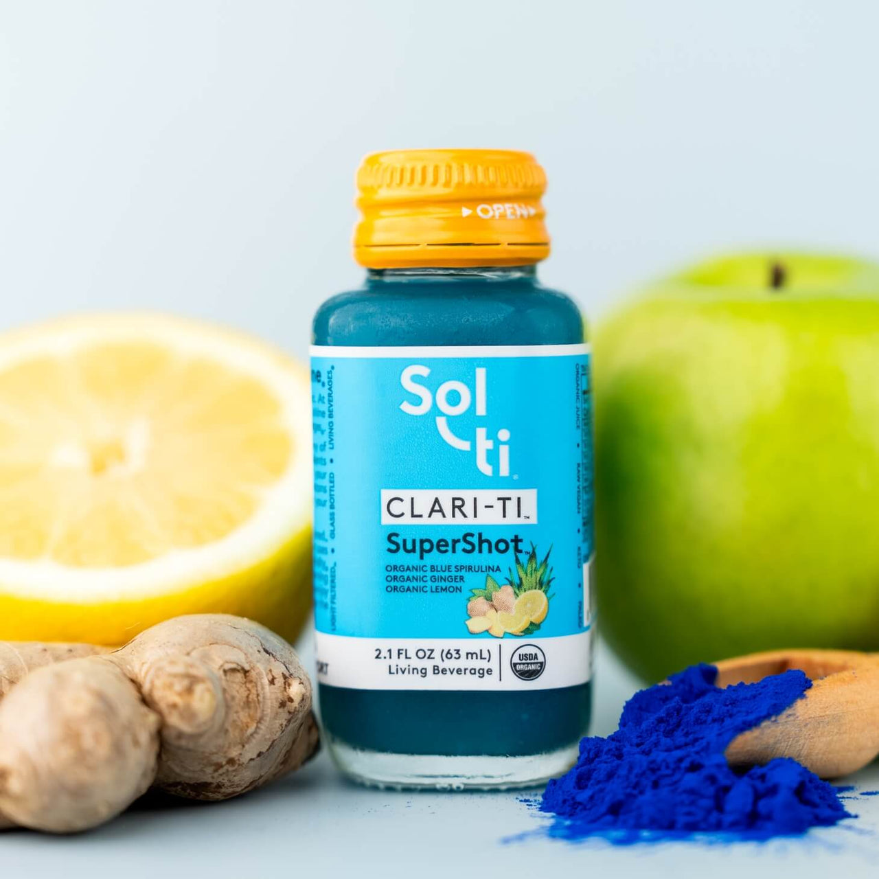 A bottle of CLARI-TI SuperShot next to blue spirulina, green apple, ginger root, and lemon slices