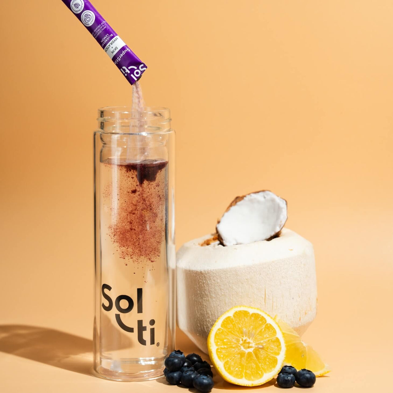 A Sol-ti Glass Bottle with Blueberry Lemon SuperStix and water, next to a young Coconut, blueberries, and lemon slices 