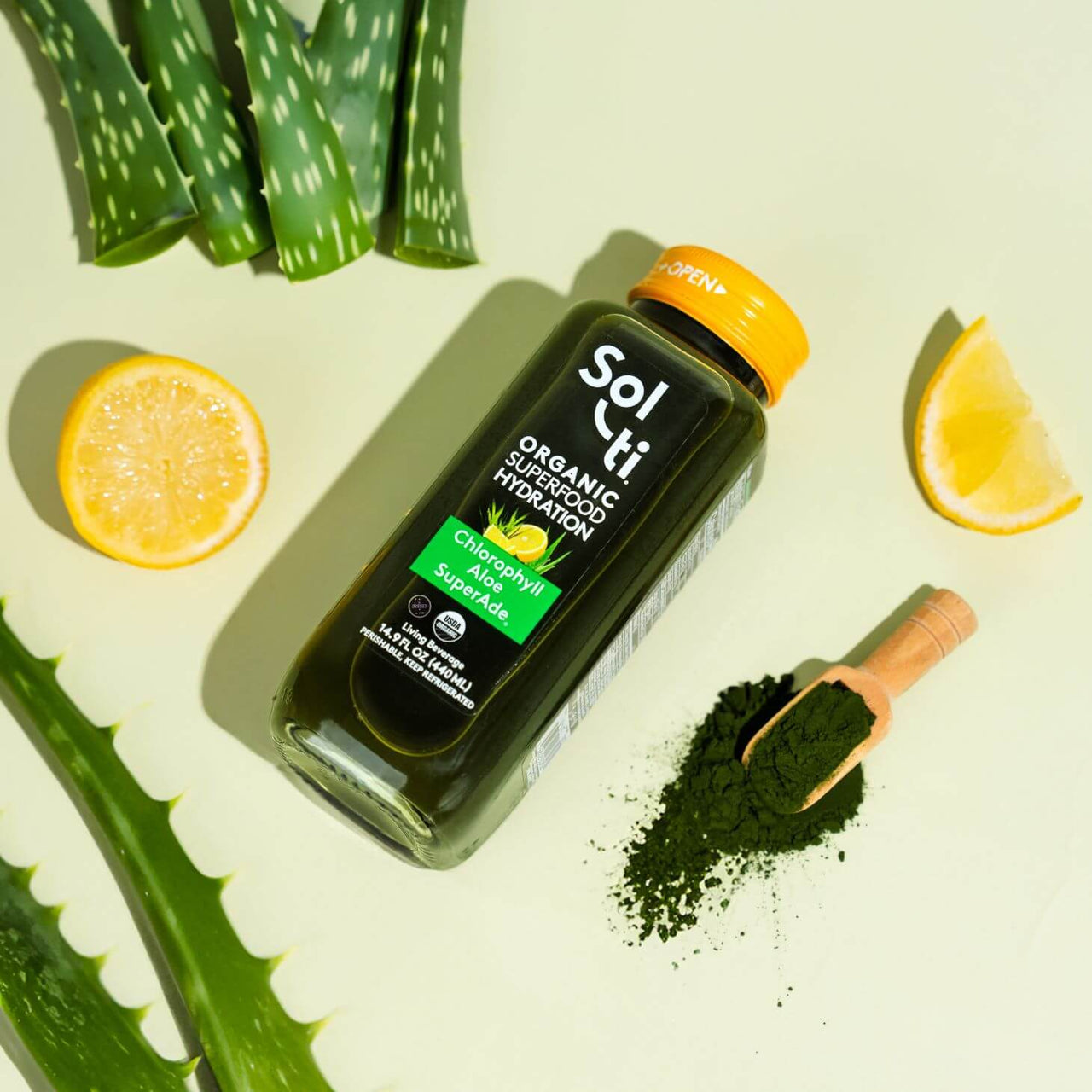 A Chlorophyll Aloe SuperAde bottle next to a lemon slices, a spoon of green spirulina, and Aloe Vera leaves