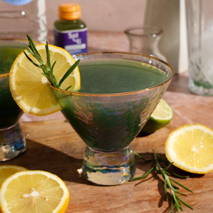 Mocktail in a martini glass with lemon and rosemary garnish on a wooden surface with Sol-ti MINDBOOST+ SuperShot in the background.
