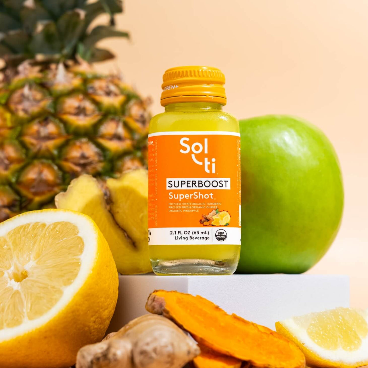 A bottle of SUPERBOOST SuperShot next to pineapple, green apple, ginger root, turmeric root, and lemon slices