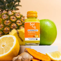 Thumbnail for A bottle of SUPERBOOST SuperShot next to pineapple, green apple, ginger root, turmeric root, and lemon slices