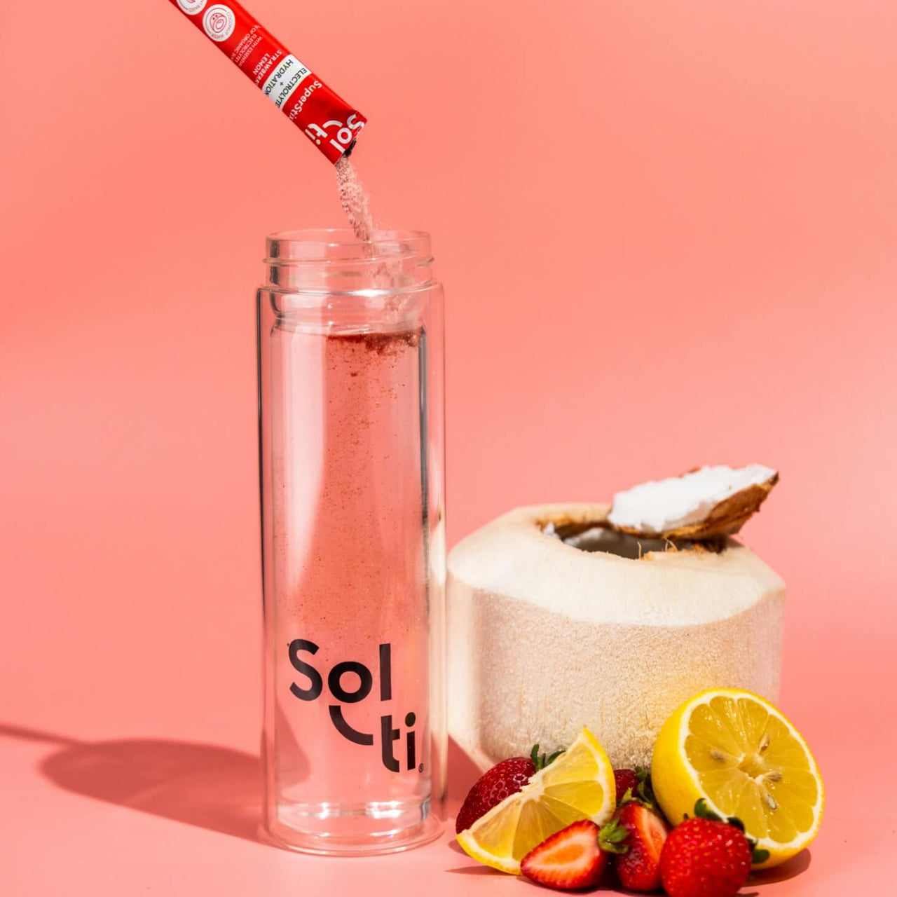 A Sol-ti Glass Bottle with Strawberry Lemon SuperStix and water, next to a young Coconut, strawberries, and lemon slices 