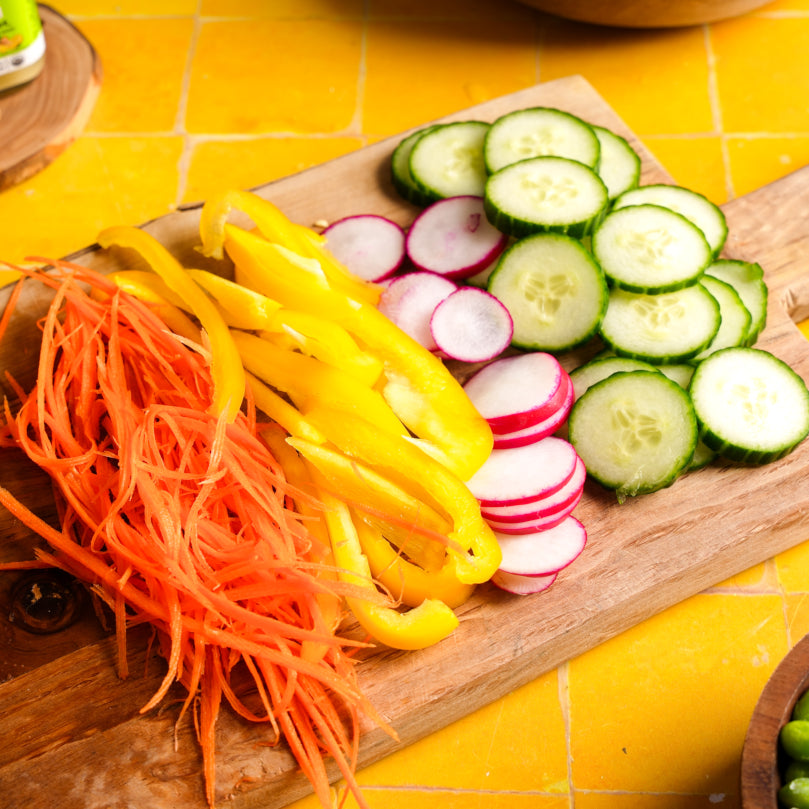 Sliced vegetables including carrots, yellow bell peppers, cucumbers, and radishes on a wooden cutting board.